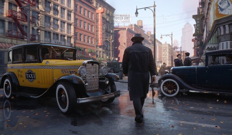 A still from the game "Mafia: Definitive Edition" showing a 1930s taxi and another dark-colored car, with a man wearing a trenchcoat and a hat, wielding a tommy gun.
